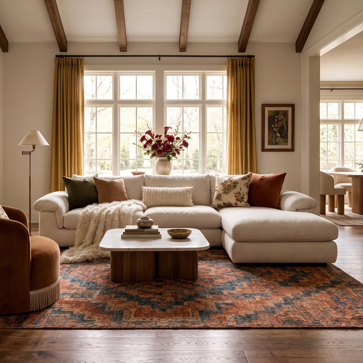 A spacious and warm living room featuring a white L-shaped sectional sofa with a chaise, styled with a velvet accent chair, decorative pillows, and a patterned rug under natural light.
